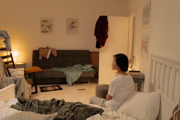 exhausted in a bed in a messy bedroom with scattered clothes and furniture.